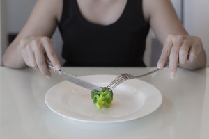 plate with one single piece of broccoli as food and woman cutting it with a fork and knife
